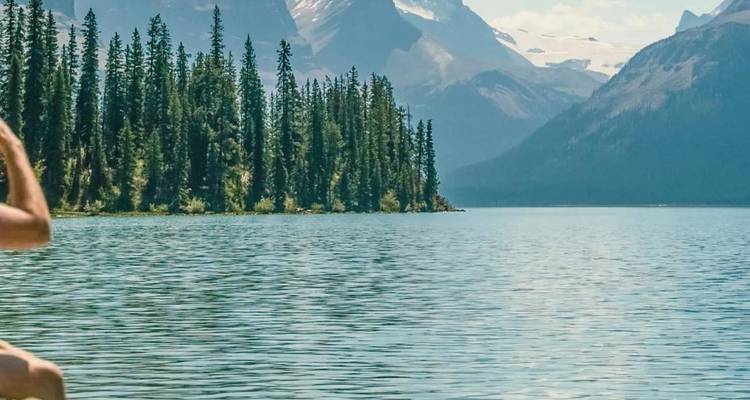 Vue partielle d'un lac alpin bordé par une forêt dense de conifères et des pics enneigés, avec une personne au bord de l'eau.