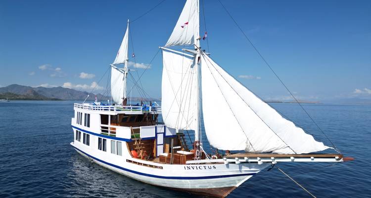 Le navire à voile à coque blanche et deux mâts 'INVICTUS' navigue sur des mers bleues calmes avec des îles arides à l'horizon.