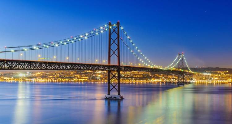 Le pont suspendu du 25 Avril illuminé enjambe le Tage la nuit avec les lumières de Lisbonne se reflétant sur l'eau.