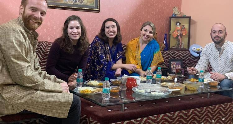 Reisende in traditioneller indischer Kleidung genießen ein hausgemachtes Abendessen auf einem Sofa um einen niedrigen Tisch.