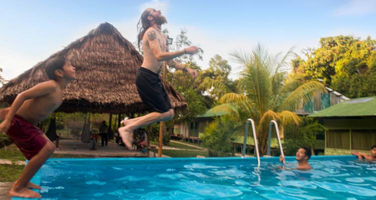 People jumping into a swimming pool with a thatched roof structure in the background.