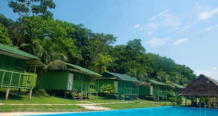 Fila de cabañas verdes junto a una piscina bajo un cielo azul.