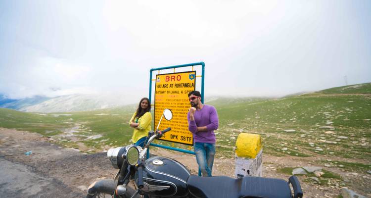 Menschen posieren neben einem Schild mit einem Motorrad in einer bergigen Region.