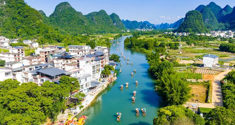 Ein Jadegrüner Fluss schlängelt sich durch ein Karsttal, das mit Bambusflößen und weißen Dörfern unter einem strahlenden Himmel in Yangshuo geprägt ist.
