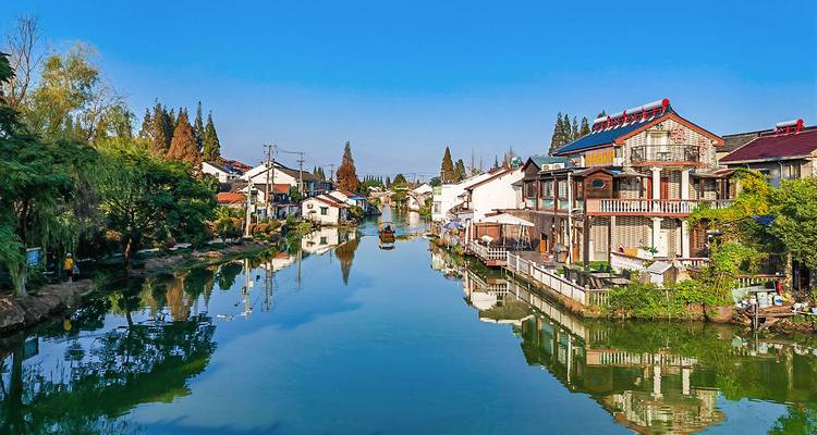 Canal paisible bordé de maisons traditionnelles blanchies à la chaux et d'arbres feuillus se reflétant dans l'eau calme de la ville lacustre de Wuzhen sous un ciel bleu.