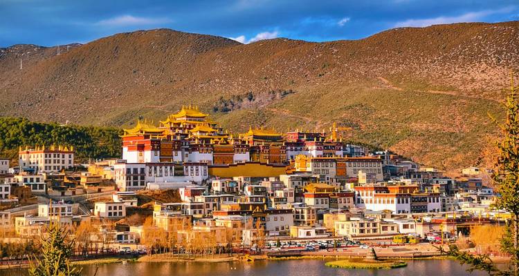 Golden-roofed Songzanlin Monastery spreads across a hillside above a reflective lake with mountains behind.
