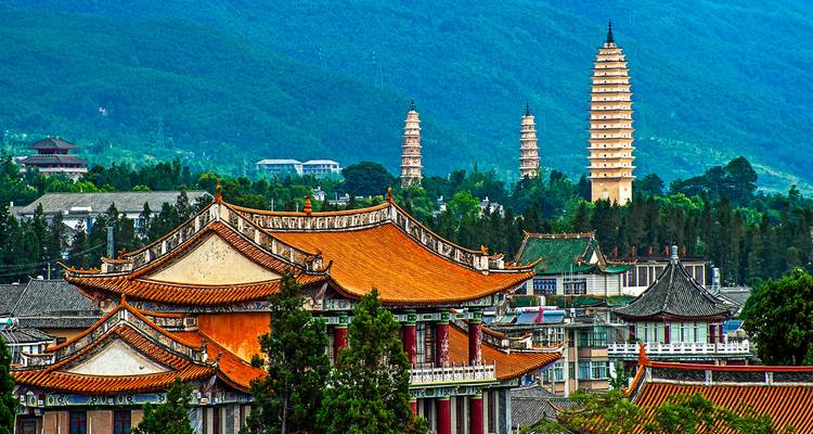 The Three Pagodas rise behind ornate temple roofs with a lush green mountain backdrop.