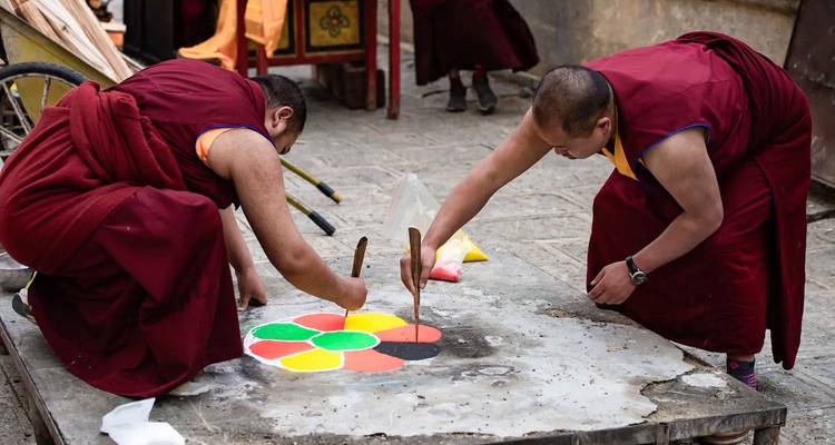 Two monks in burgundy robes meticulously create a colorful sand mandala on a stone platform.