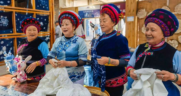 Four smiling women in bright ethnic costumes demonstrate traditional tie-dye techniques inside a workshop.