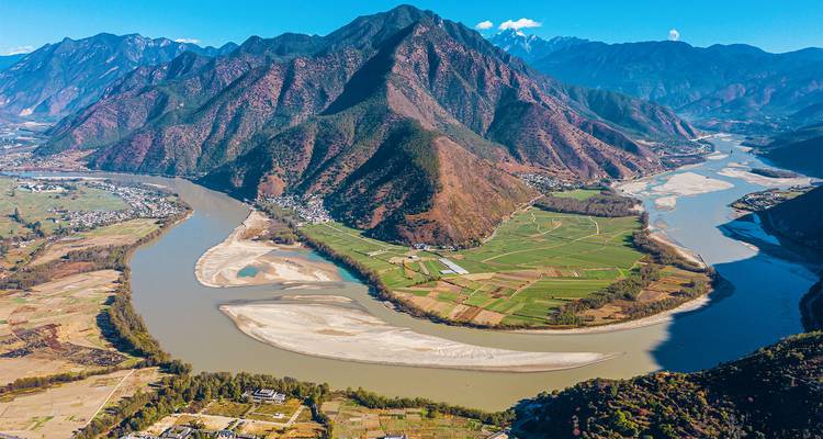 An aerial panorama shows the sweeping first bend of the Yangtze River around green farmland and a massive mountain.