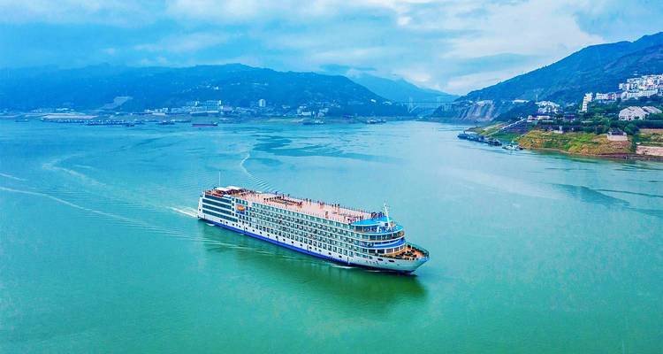 Grand navire de croisière fluviale multi-étages naviguant à travers une large section verdoyante du Yangtsé avec des montagnes brumeuses alentour.