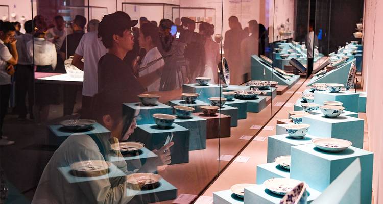 Bezoekers bestuderen porseleinartefacten die op turquoise standaards achter glas in een moderne museumgalerij zijn uitgestald.