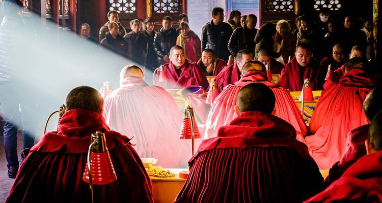 Des moines vêtus de robes rouges chantent à l'intérieur d'une salle de temple obscure tandis qu'un rayon de lumière dramatique illumine la scène.