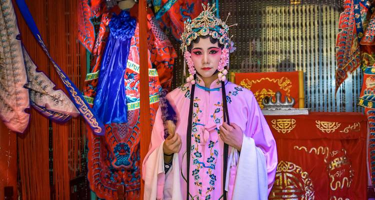 Aufwendig kostümierter chinesischer Operndarsteller in einem reich verzierten Backstage-Bereich, der einen Fächer in der Hand hält.