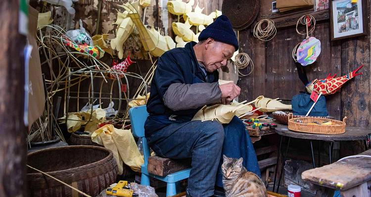 Een ambachtsman maakt traditionele papieren figuren in een rustieke werkplaats vol met materialen terwijl een kat ergens in de buurt zit.