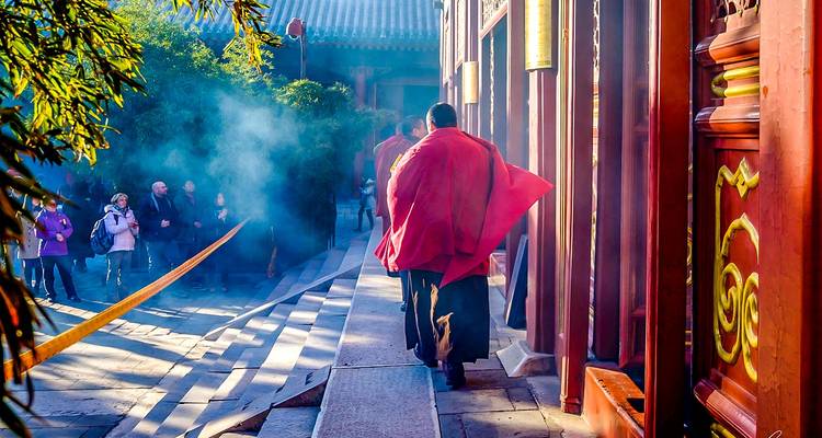 Un monje envuelto en una túnica roja camina por un patio lleno de incienso mientras los visitantes observan en el templo.