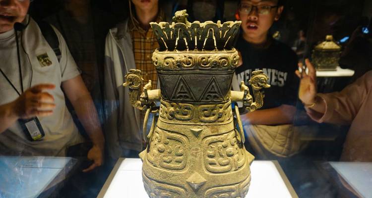 Besucher betrachten ein kunstvoll geschnitztes antikes Bronzegefäß, das unter Spotlicht in einer dunklen Museumsgalerie ausgestellt ist.