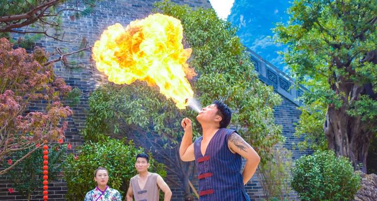 Ein Performer stößt einen Feuerstrahl in die Luft aus, während Zuschauer in einem von Grünflächern und Steinmauern umrahmten Innenhof zuschauen.