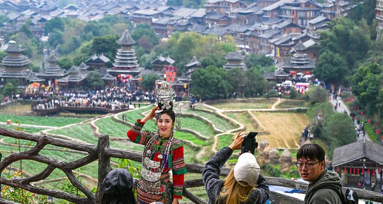 Eine Frau in aufwendiger Miao-Tracht posiert auf einem Zaun mit Blick auf Reisterrassen und ein traditionelles Dorf.