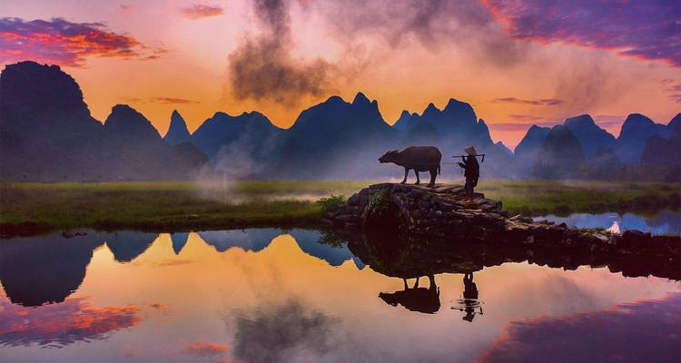 Silhouettierter Landwirt mit Wasserbüffel in einer nebligen Karstlandschaft, perfekt in stillem Wasser bei Sonnenaufgang gespiegelt.