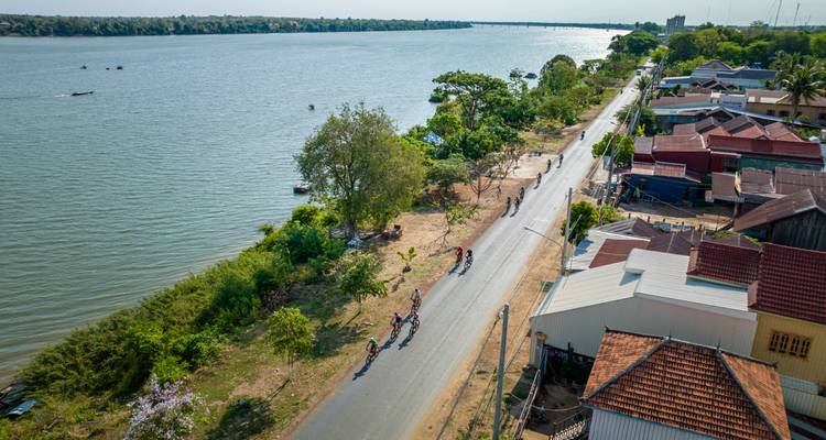 Cyclists on a road by a river with a distant cityscape.