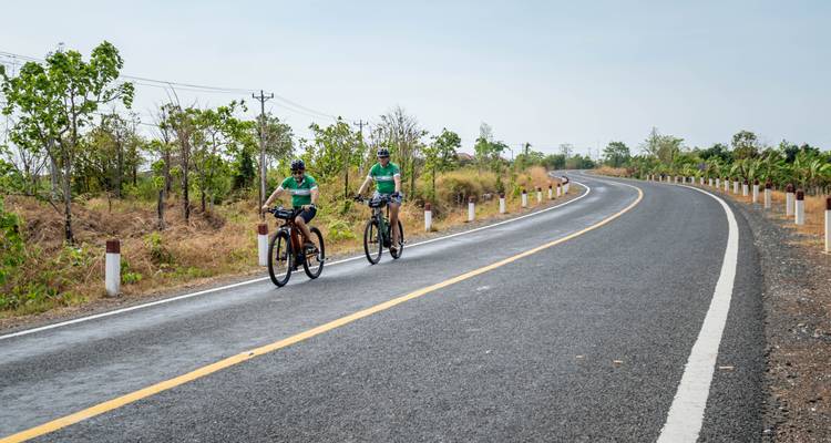 Two cyclists on a rural road lined with pylons and dry vegetation.