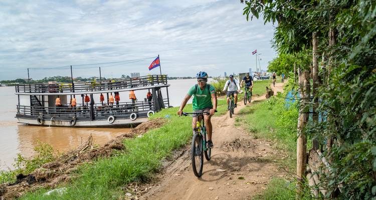 Cyclists on a dirt path beside a river with a boat nearby, waving Cambodian flag