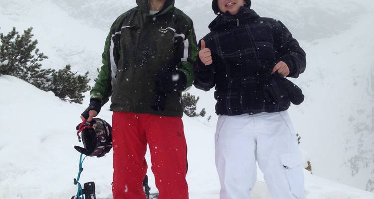 Deux personnes debout en équipement de neige avec des montagnes enneigées en arrière-plan.