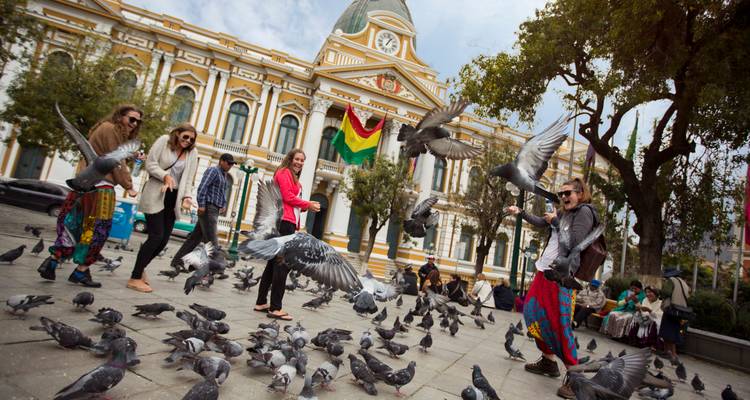 Voyageurs jouant parmi les pigeons devant un bâtiment gouvernemental orné avec le drapeau bolivien