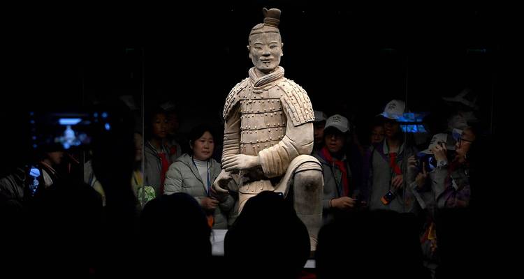 Single illuminated terracotta warrior statue displayed in a dark museum gallery, surrounded by visiting tourists behind glass.