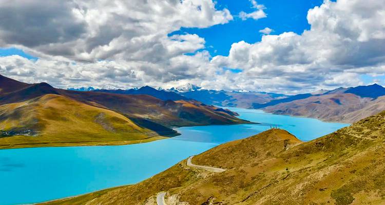Panoramic view of turquoise Yamdroktso Lake winding between brown hills and distant snow peaks under dramatic clouds.