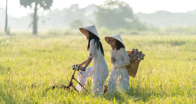 Dos mujeres con vestimenta tradicional en un campo con una bicicleta y una canasta de frutas.
