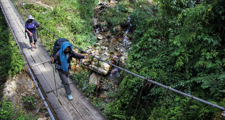 Trekking path with two hikers crossing a suspension bridge over a river.