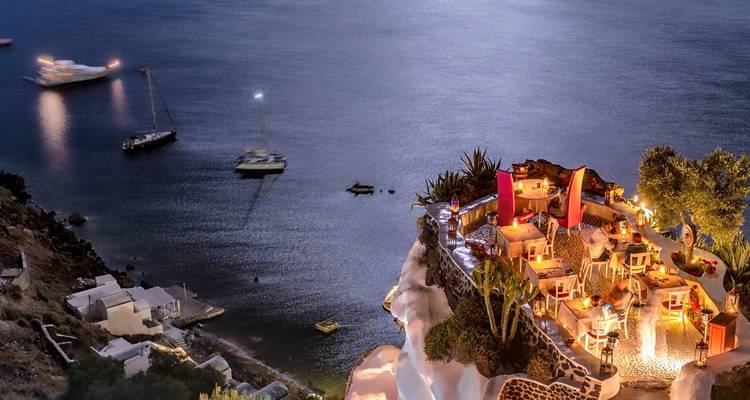 Vue d'un restaurant illuminé à flanc de falaise avec la mer et des bateaux la nuit.