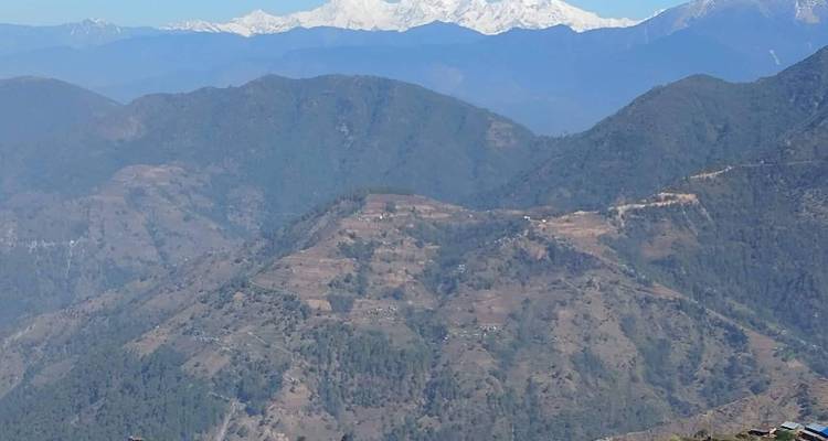 Les lointains pics enneigés de l'Himalaya se dressent au-dessus des collines ondulantes vertes et brunes sous un ciel dégagé.