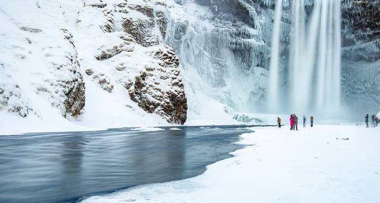 La cascada congelada Skógafoss desciende junto a acantilados cubiertos de hielo mientras los visitantes exploran la orilla nevada del río.