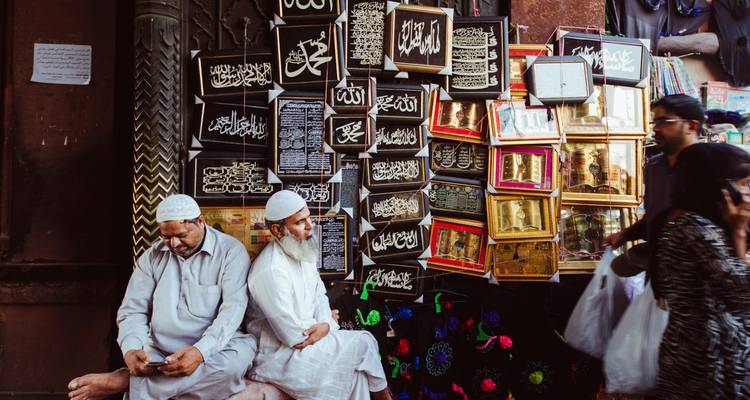 Deux hommes âgés en bonnets blancs sont assis parmi des œuvres d'art de calligraphie arabe encadrées dans un marché animé de Delhi.