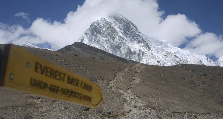 Geel trailbord dat naar Everest Base Camp wijst, met op de achtergrond een hoge sneeuwbedekte piek en blauwe lucht.
