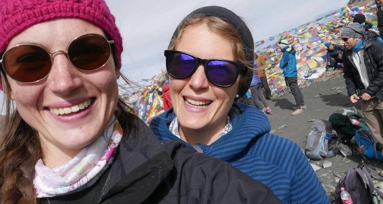 Deux trekkeurs souriants prenant un selfie à un col de montagne élevé avec des drapeaux de prière colorés en arrière-plan