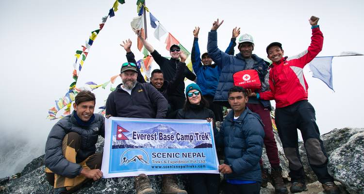 Trekkers celebrate with flags and a banner on a rocky summit in cloudy conditions.