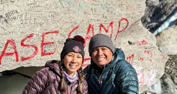 Close portrait of two trekkers smiling beside the Base Camp rock with red lettering.