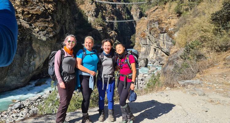 Four trekkers stand on a Himalayan trail near a river with suspension bridges overhead.