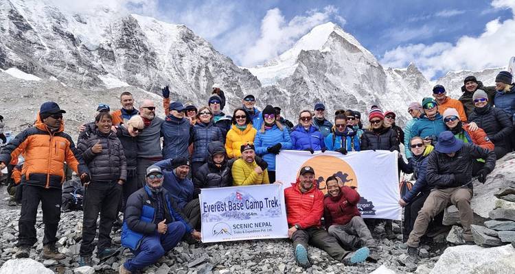 Large group celebrates at Everest Base Camp with snowy peaks behind them holding banners.