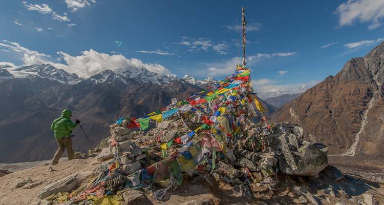 Randonneu se tenant debout à côté de drapeaux de prière colorés sur un sommet de montagne avec des montagnes enneigées en arrière-plan.