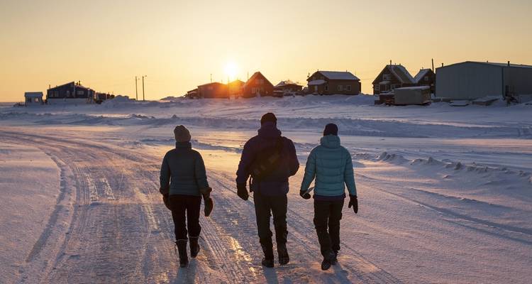Tres personas caminando por una carretera nevada hacia la puesta de sol.