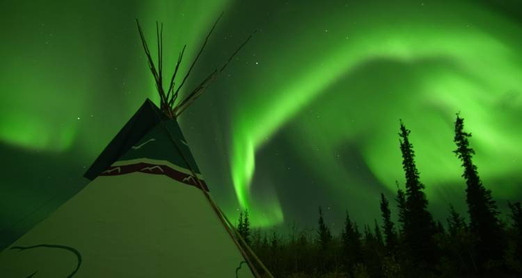 Northern lights above a tipi with a forest silhouette.