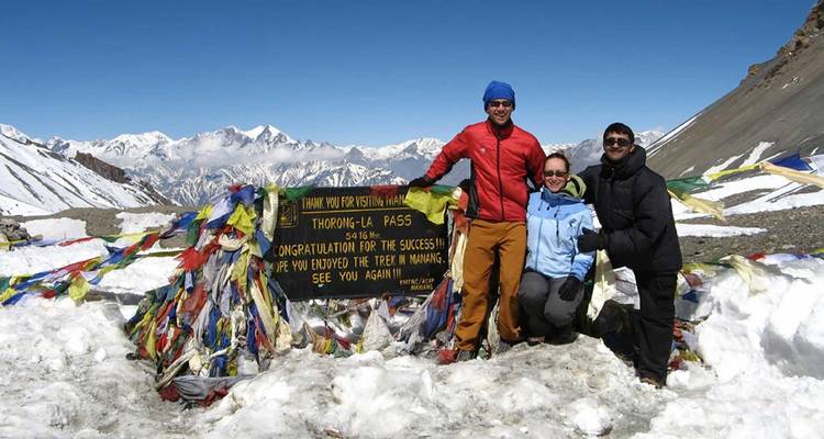 Three people posing at Thorong La Pass with prayer flags.