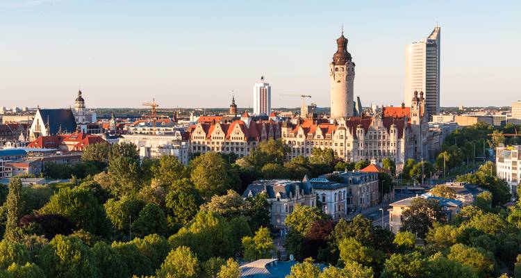 Panoramisch uitzicht op Leipzig met de historische Nieuwe Stadhuistoren en moderne City-Hochhaus die boven rooddakige gebouwen uitrijzen, omlijst door weelderig groen onder helder avondlicht.