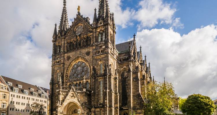 Gotische kerk met sierlijke torenspitsen en roosvenster tegen een gedeeltelijk bewolkte blauwe hemel, omringd door stadsgebouwen.
