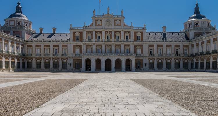 Fachada de palacio real simétrica frente a un amplio patio de piedra en un día despejado.